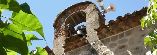Campanario de piedra y ladrillo de Santa Cruz del Valle, con una campana bajo un arco y rodeado de vegetación.